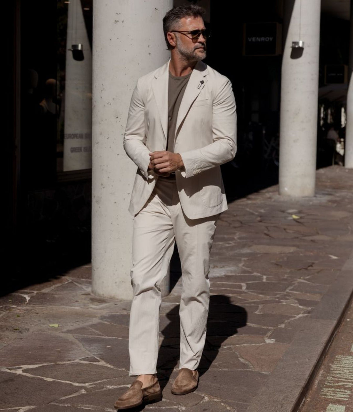 A man wearing a light beige two-piece suit with a brown t-shirt and brown suede loafers, standing on a stone sidewalk in front of a building with white columns.