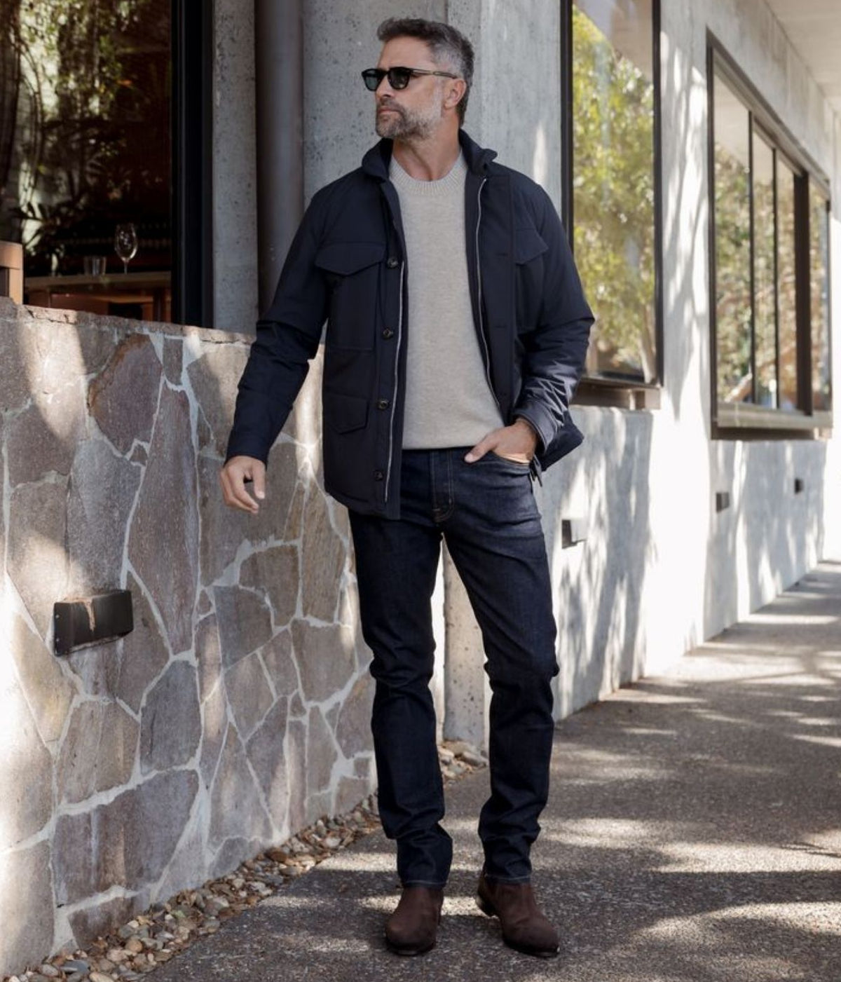 A man wearing a dark navy utility jacket over a light beige crewneck sweater, dark jeans, and brown boots, standing outdoors next to a stone wall and modern building.