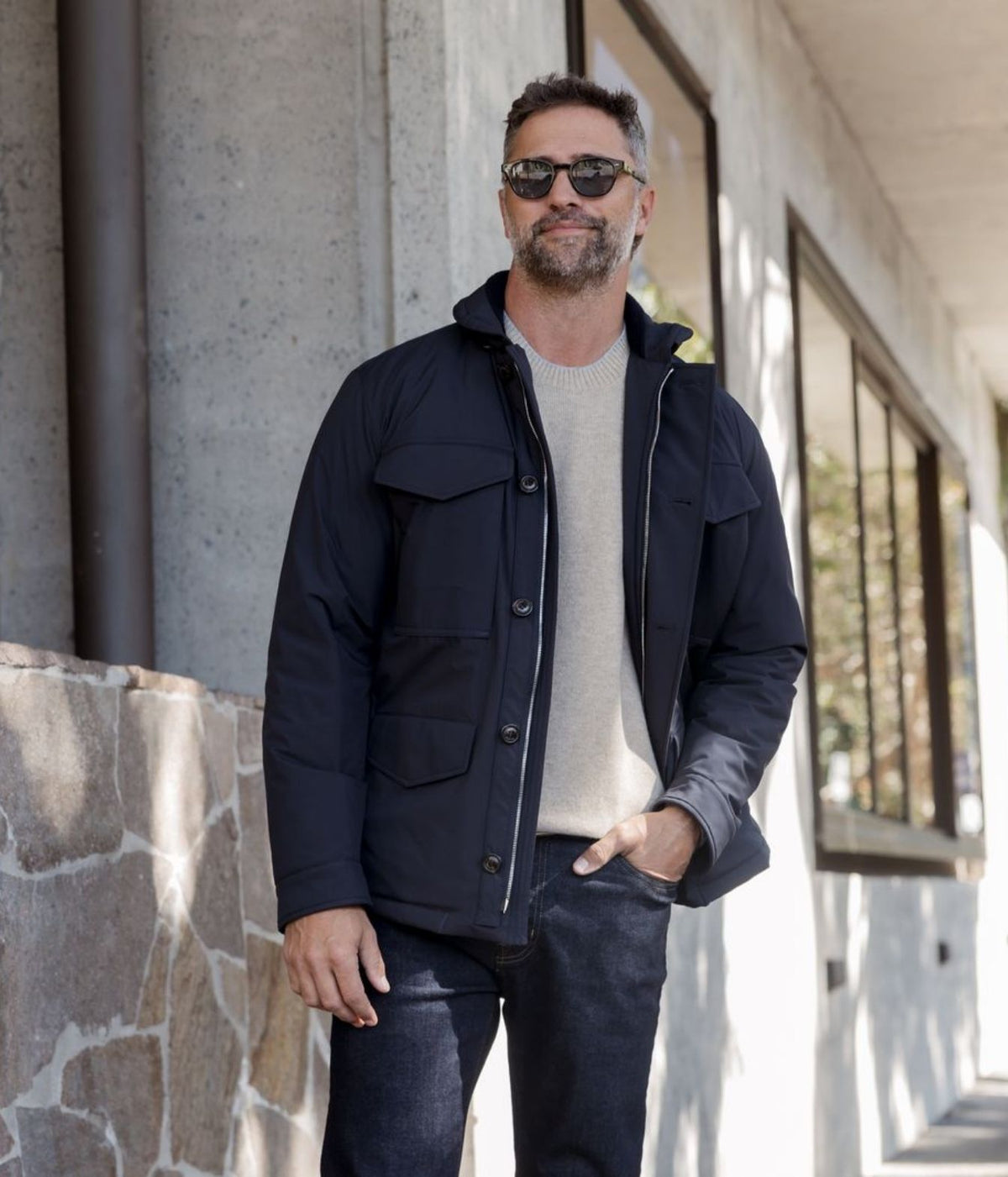A man wearing a dark navy blue utility jacket over a light beige knit sweater and dark jeans, standing outdoors in front of a modern concrete building.