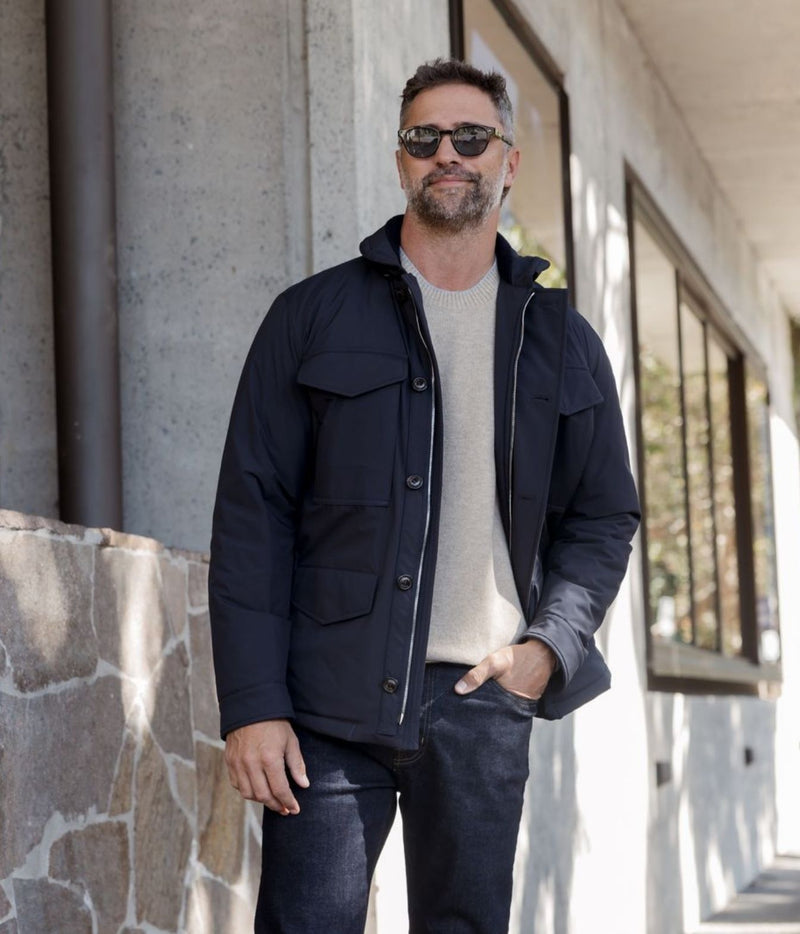 A man wearing a dark navy blue utility jacket over a light beige knit sweater and dark jeans, standing outdoors in front of a modern concrete building.