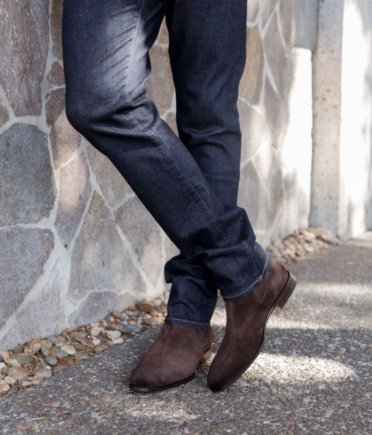 A person wearing dark brown suede Chelsea-style boots with dark blue jeans, standing on a gravel path next to a stone wall.