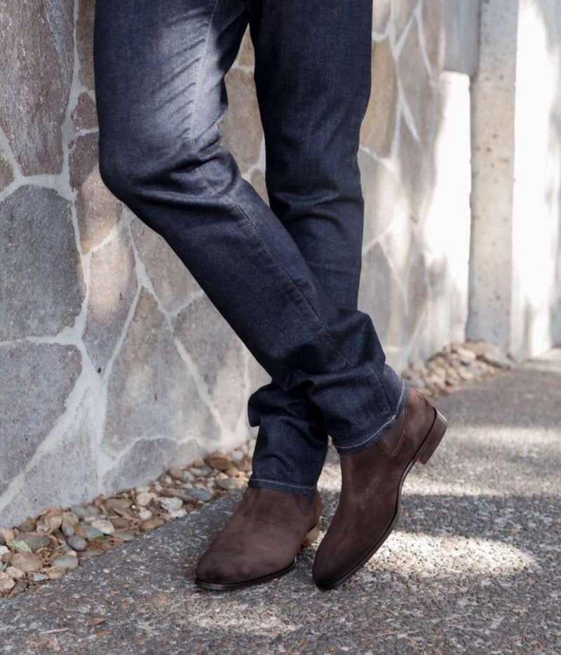 A person wearing dark brown suede Chelsea-style boots with dark blue jeans, standing on a gravel path next to a stone wall.