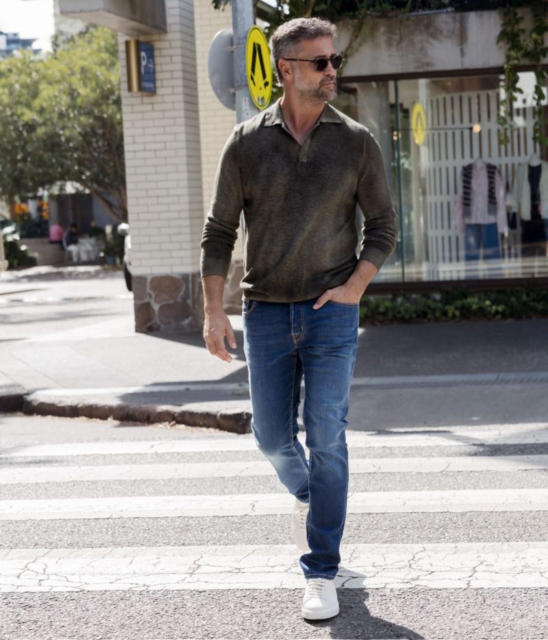 A man wearing a dark olive green long-sleeve polo shirt, blue jeans, and white sneakers walks across a city crosswalk.