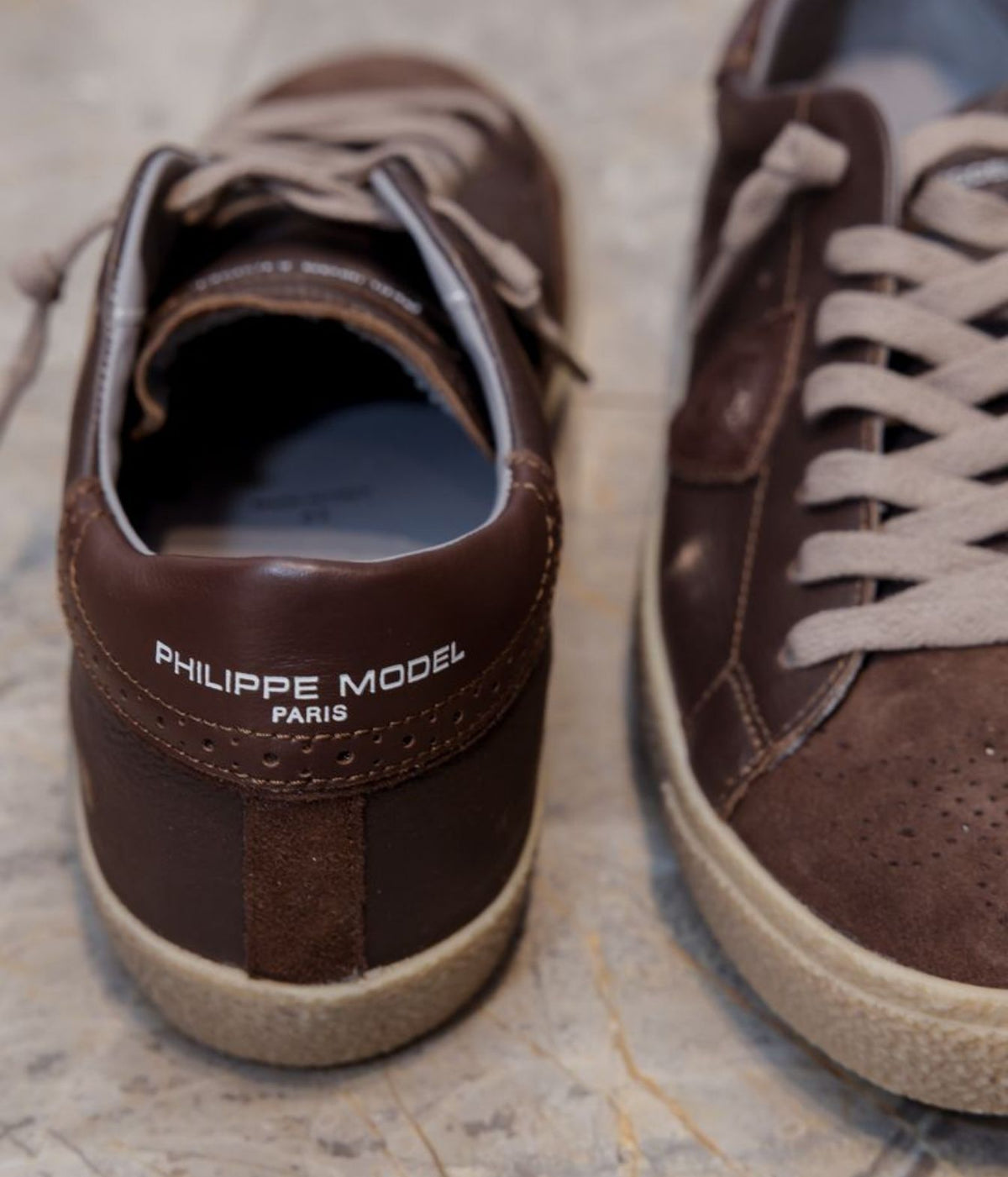 A pair of dark brown leather low-top sneakers with beige laces and beige rubber soles, featuring the text 'PHILIPPE MODEL PARIS' on the heel of the left shoe, shown on a marble surface.