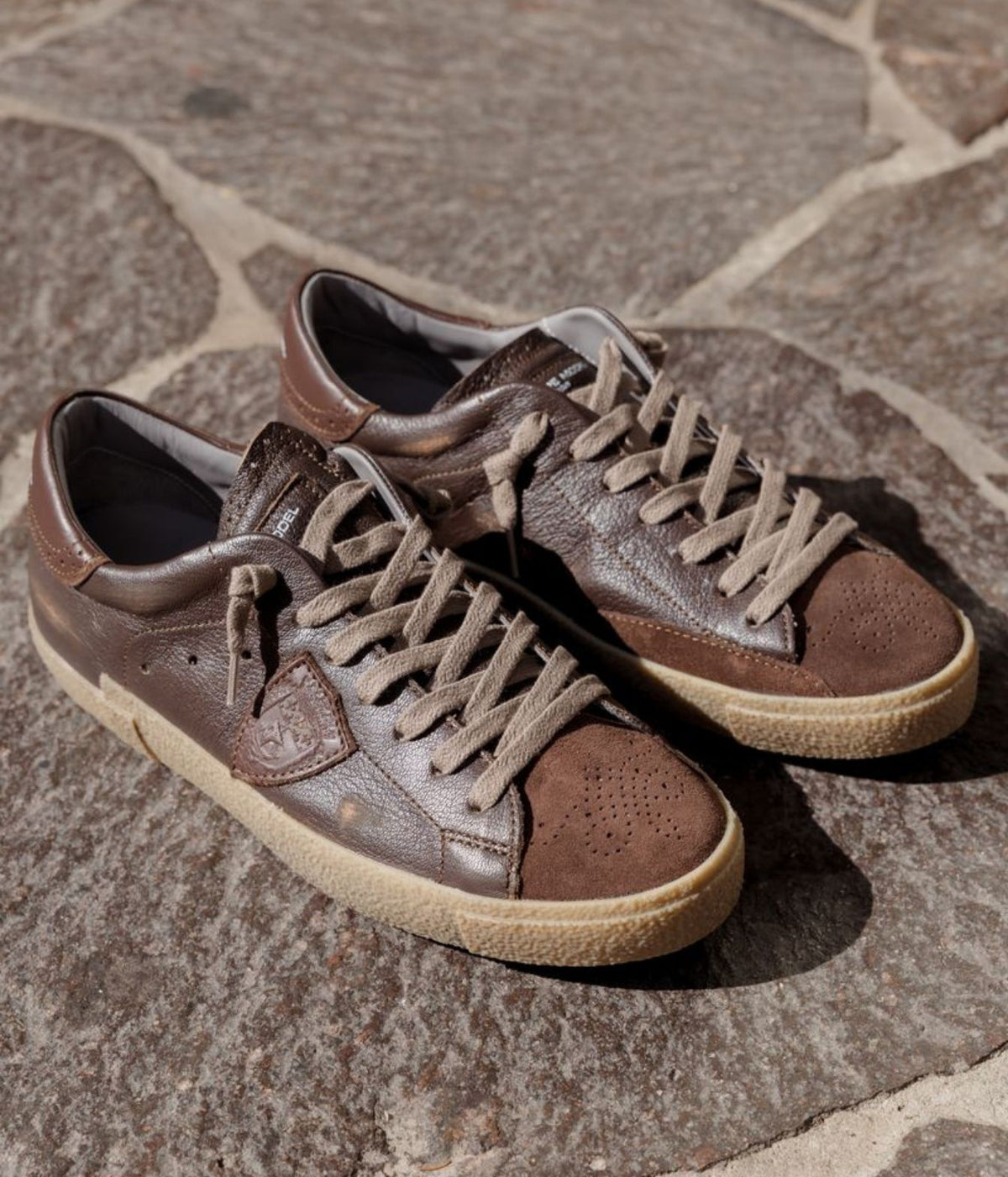 A pair of brown leather low-top sneakers with beige laces and beige rubber soles, photographed on a stone pavement background.