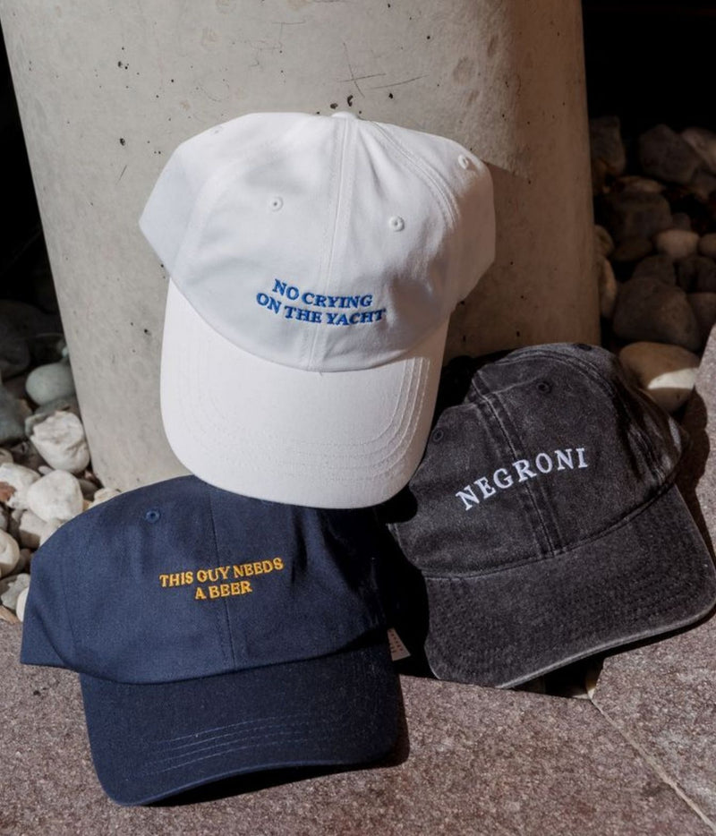 Three embroidered baseball caps arranged on stone pavement in front of a concrete column: a white cap with blue text reading 'NO CRYING ON THE YACHT,' a navy cap with yellow text reading 'THIS OLY NEEDS A BEER,' and a dark charcoal cap with white text reading 'NEGRONI.'.