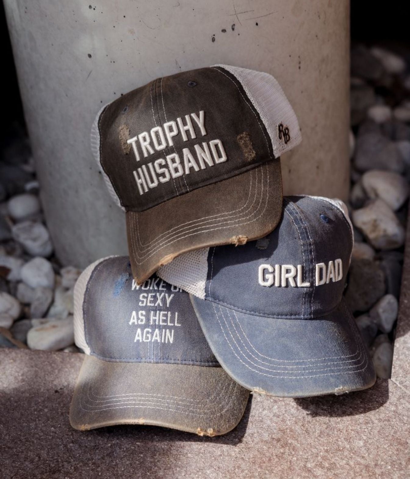 Three distressed trucker-style baseball caps stacked together outdoors near a concrete post and gravel. The top cap is dark brown with white text reading 'TROPHY HUSBAND' and mesh side panels.