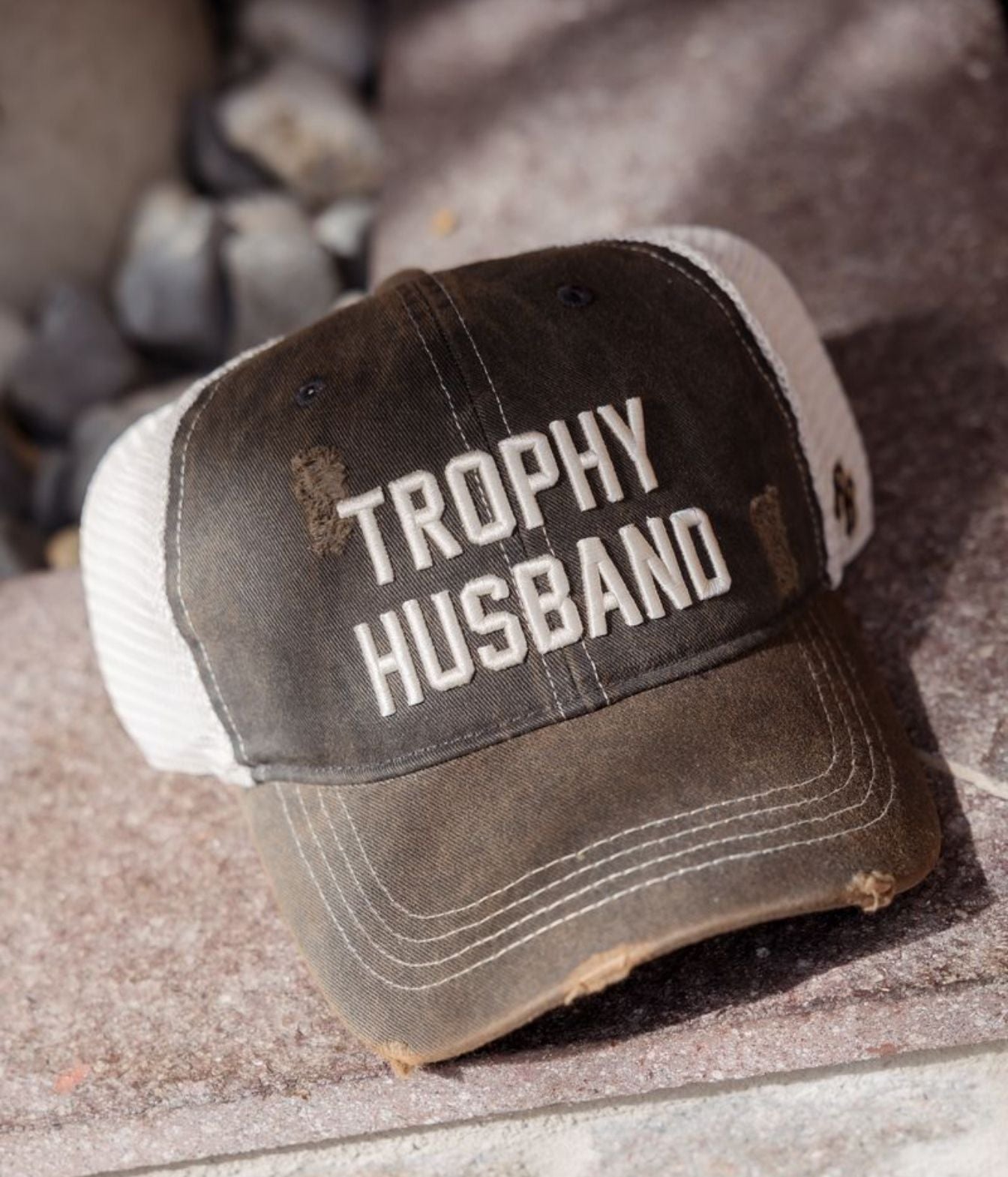 A distressed trucker-style baseball cap with a dark brown front panel and white mesh back, featuring the words 'TROPHY HUSBAND' embroidered in white on the front. The cap is resting on a stone surface with blurred rocks in the background.