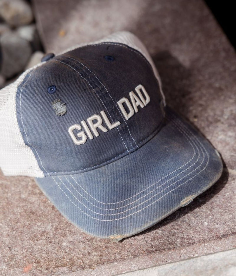 A distressed trucker hat with a dark blue front panel and white mesh back, featuring the embroidered text 'GIRL DAD' on the front. The hat is resting on a textured stone surface.