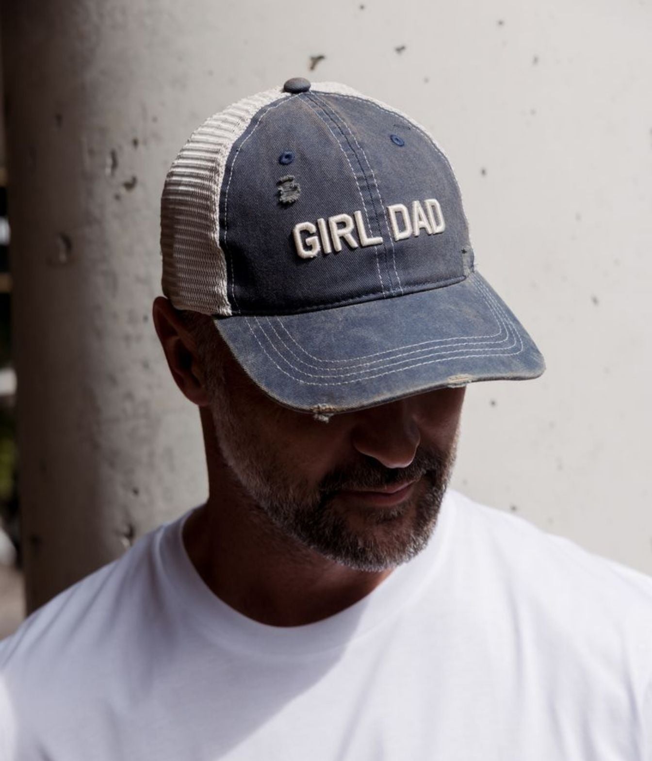 A man wearing a distressed trucker-style baseball cap with a blue front panel and mesh back, featuring the words 'GIRL DAD' embroidered in white on the front. He is wearing a white t-shirt and looking down, set against a concrete pillar background.