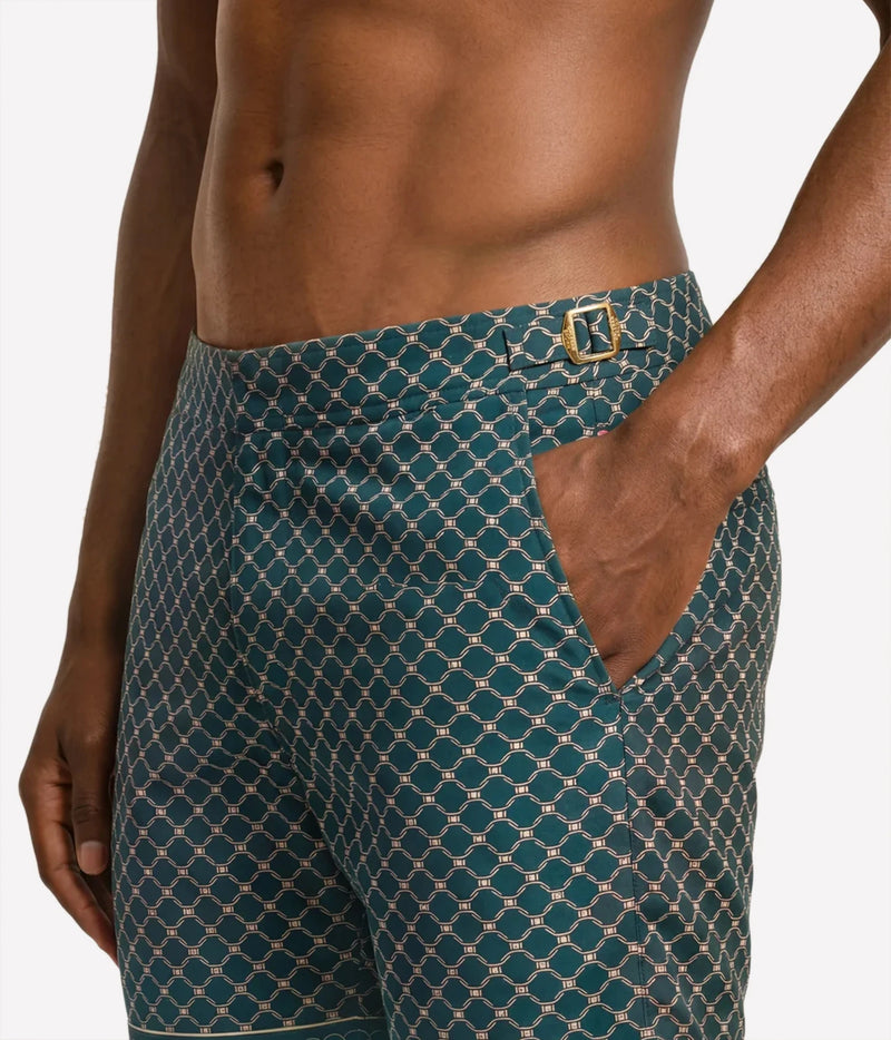 A close-up of a male model wearing teal and gold patterned swim trunks against a light gray background.