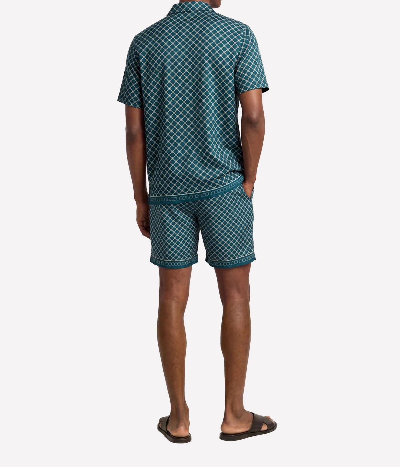 A man viewed from the back wearing a matching short-sleeve collared shirt and shorts set in a dark teal green and white geometric pattern, paired with black slide sandals, against a light gray background.