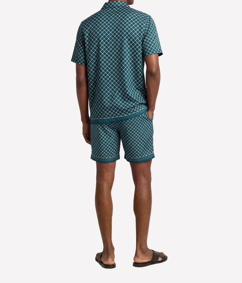 A man viewed from the back wearing a matching short-sleeve collared shirt and shorts set in a dark teal green and white geometric pattern, paired with black slide sandals, against a light gray background.