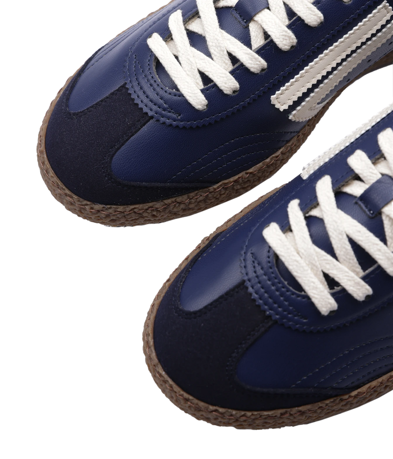 Close-up of a pair of low-top sneakers with navy blue and dark navy panels, white laces, and brown braided soles on a white background.