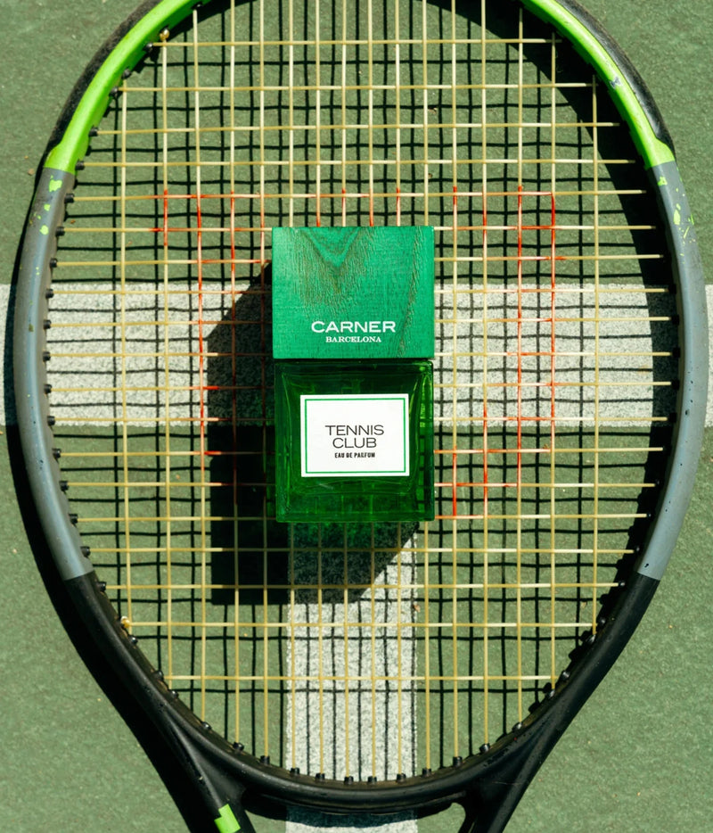 A green rectangular perfume box and matching bottle labeled CARNER TENNIS CLUB are placed on the strings of a tennis racket on a tennis court.