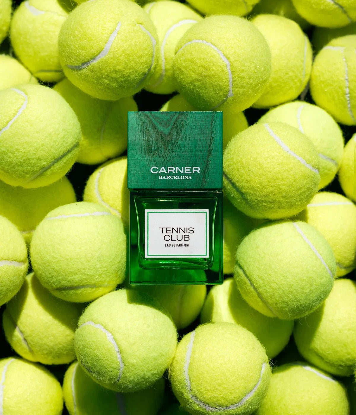 A green rectangular perfume bottle labeled 'CARNER BARCELONA TENNIS CLUB' sits centered on a dense pile of bright yellow-green tennis balls. The bottle label reads 'TENNIS CLUB' and 'EAU DE PARFUM'.