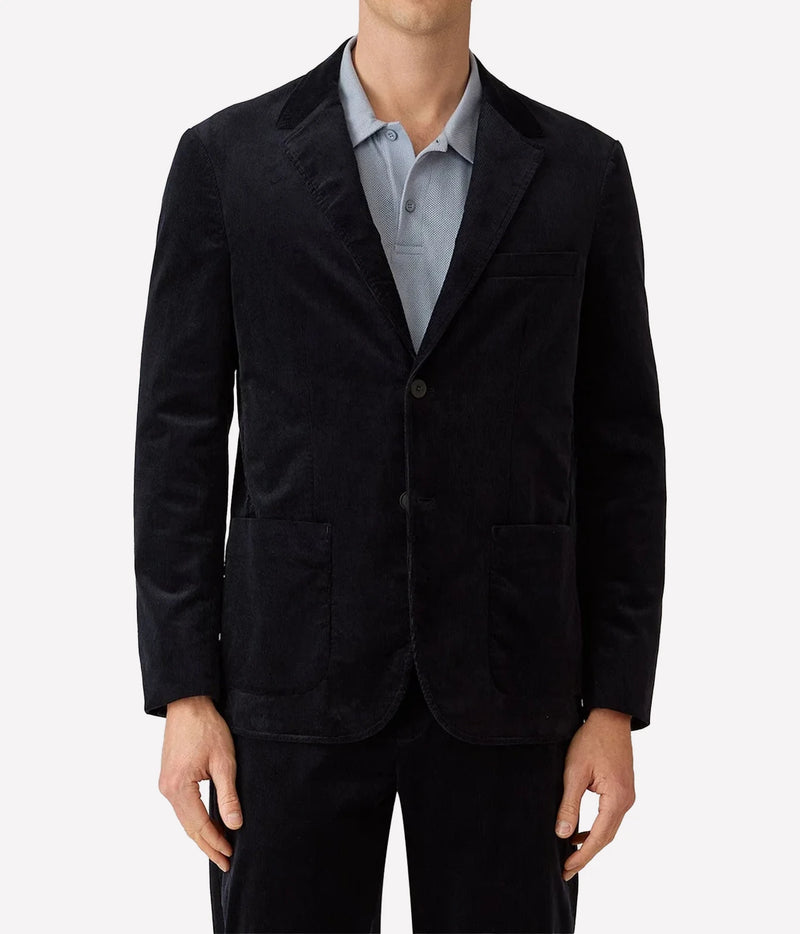 A man wearing a black corduroy blazer over a light blue collared shirt, shown from the neck down against a plain light gray background.