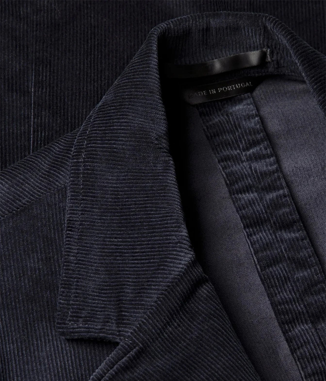 Close-up of a dark navy corduroy jacket with a visible collar and inner lining, showing a small black neck label that reads 'MADE IN PORTUGAL'.