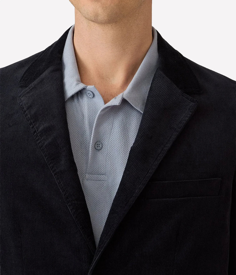 Close-up of a person wearing a dark corduroy blazer over a light blue textured polo shirt, against a plain light background.
