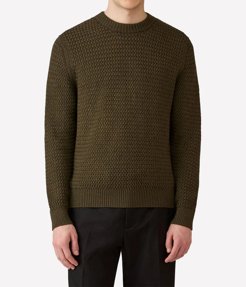 A man wearing a textured olive green crewneck sweater and black trousers, shown from the neck down against a light gray background.