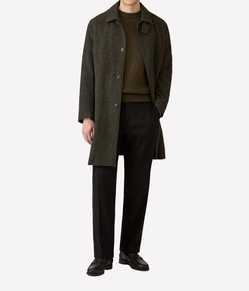 A model wearing a dark olive green wool-blend overcoat over a textured olive green sweater, black trousers, and black leather shoes, photographed against a solid light gray background.
