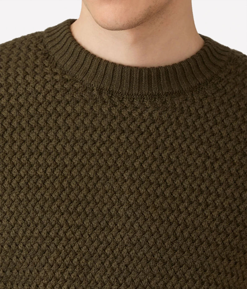 Close-up of a person wearing a textured knit crewneck sweater in olive green, showing the ribbed collar and fine knit pattern.