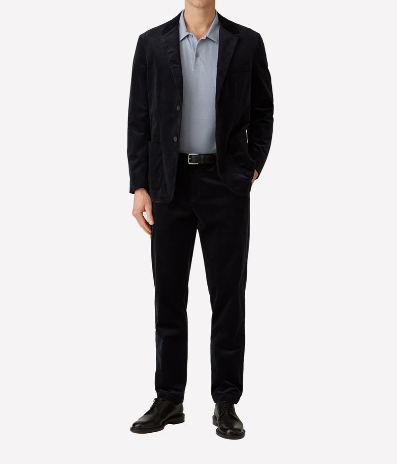 A man wearing a dark navy corduroy blazer and matching trousers, styled with a light blue polo shirt, black belt, and black leather dress shoes, against a plain white background.