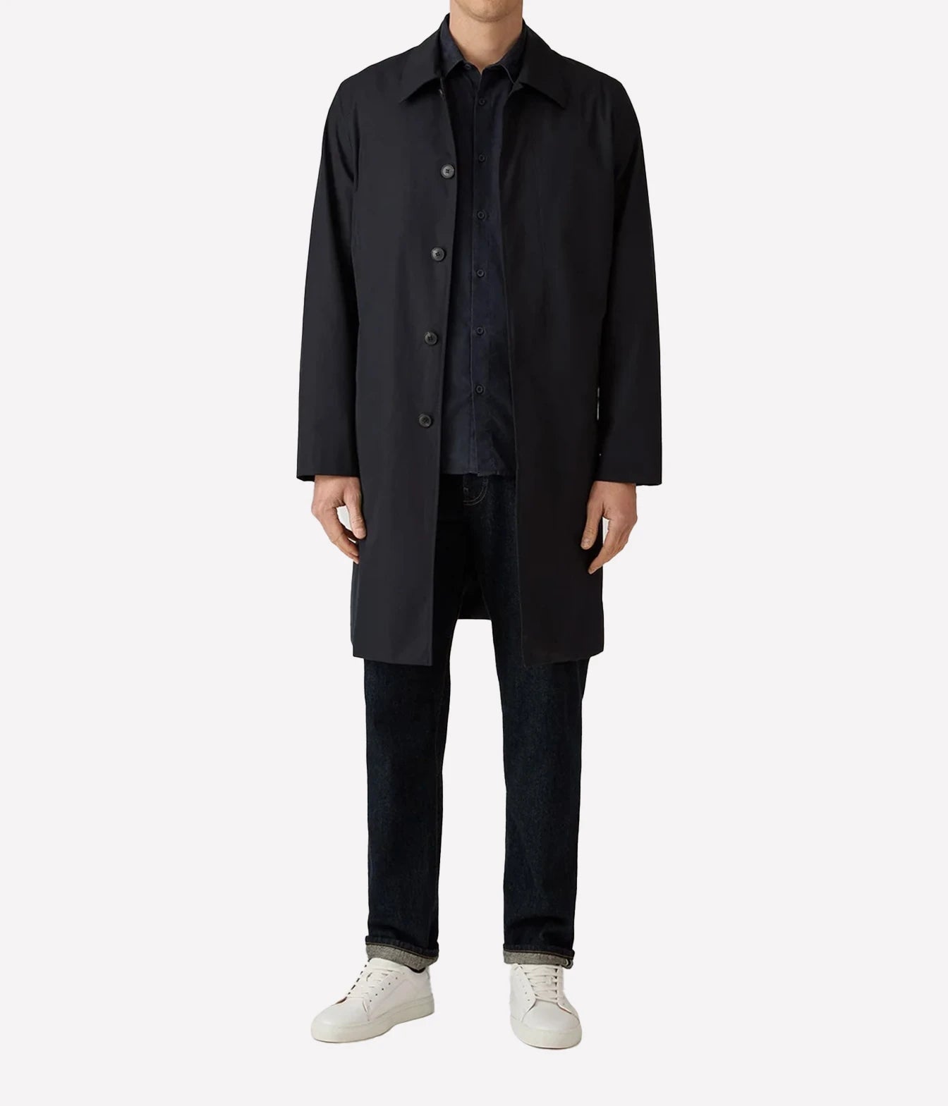 A model wearing a long black coat over a dark shirt and jeans, paired with white sneakers, shot against a plain light gray background.