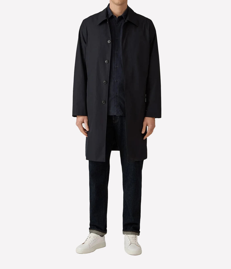 A model wearing a long black coat over a dark shirt and jeans, paired with white sneakers, shot against a plain light gray background.