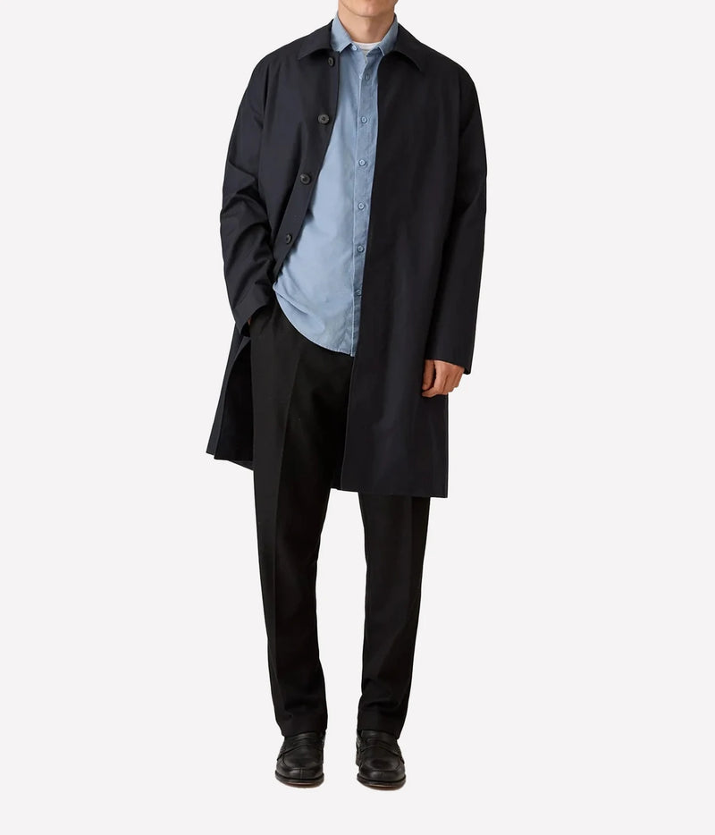 A model wearing a dark navy blue trench coat over a light blue button-down shirt, black trousers, and black shoes, photographed against a solid light gray background.