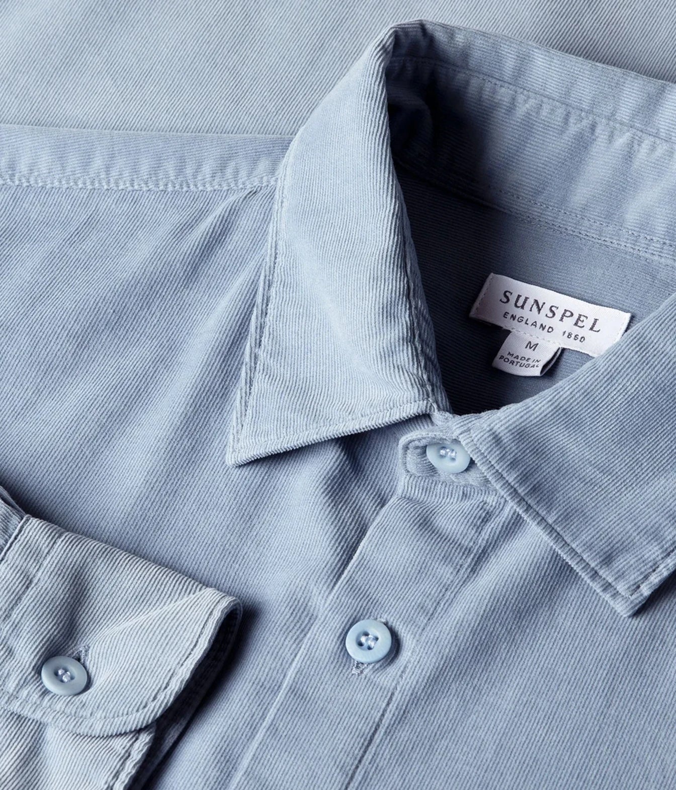 Close-up of a light blue corduroy shirt collar with a visible Sunspel label, showing the fabric texture and button details.