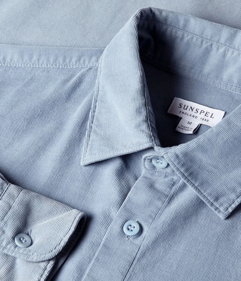 Close-up of a light blue corduroy shirt collar with a visible Sunspel label, showing the fabric texture and button details.