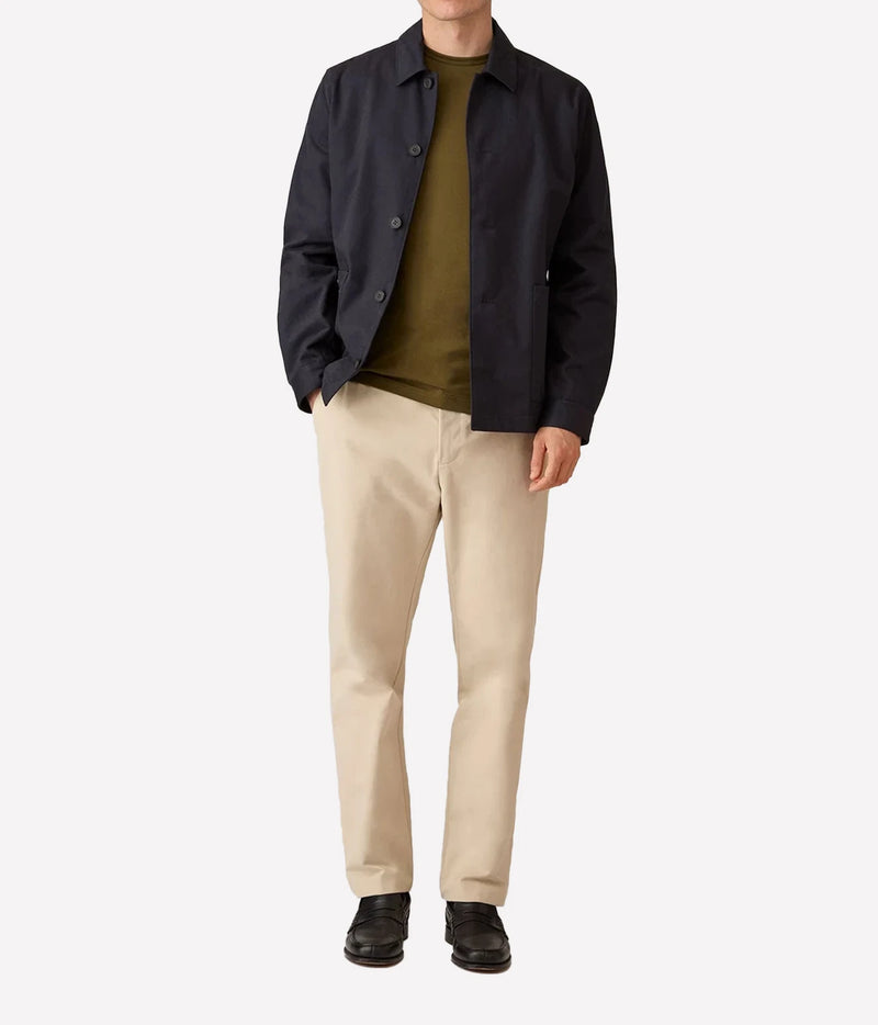 A model wearing a dark navy blue jacket over an olive green t-shirt, paired with beige trousers and black shoes, photographed against a solid light gray background.