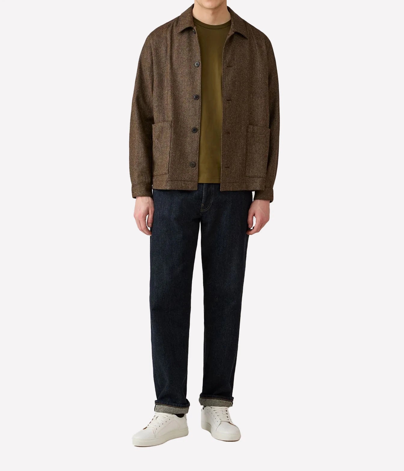 A model wearing a brown textured button-up jacket over an olive green t-shirt, dark blue jeans, and white sneakers, photographed against a solid light gray background.